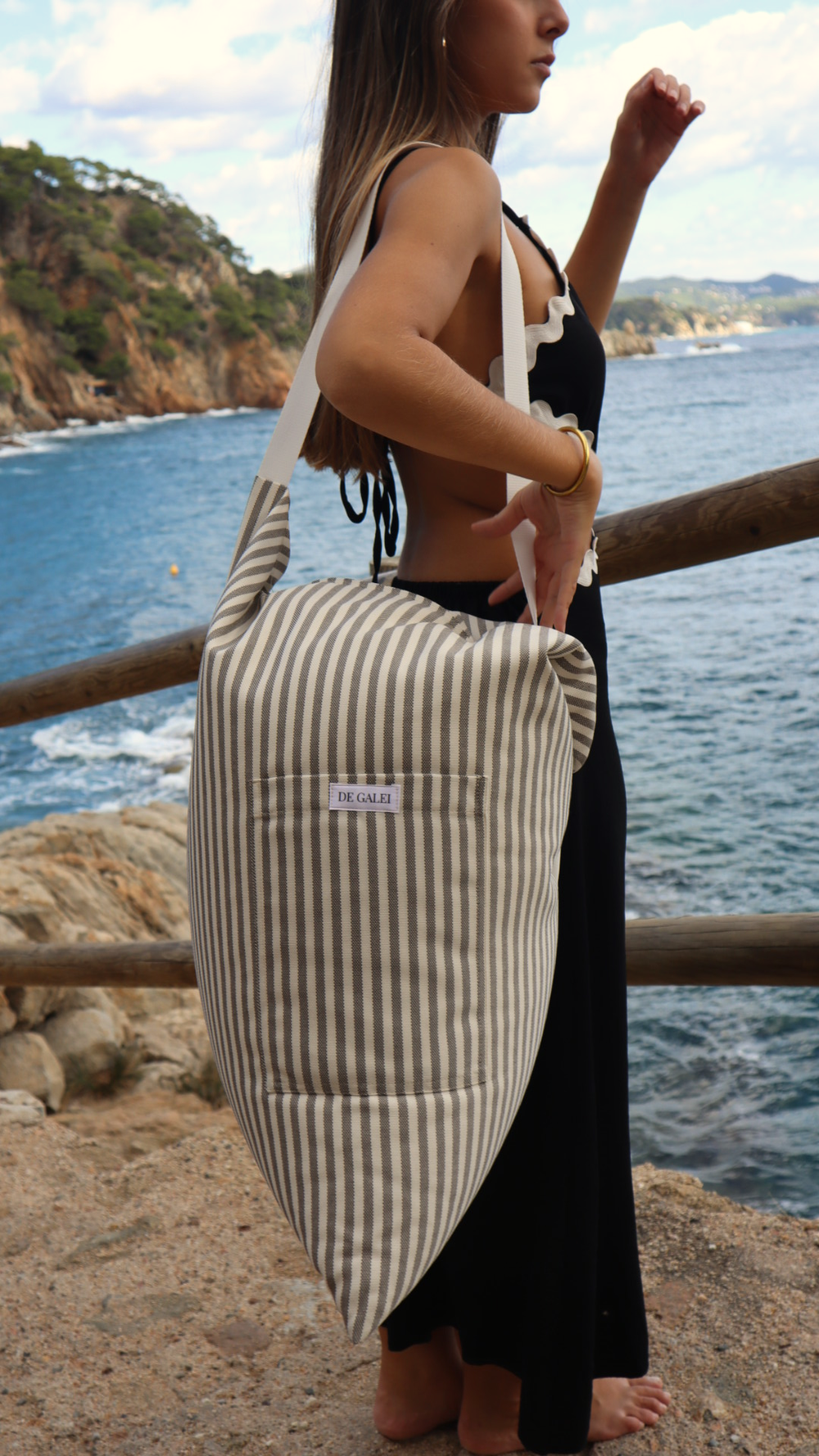 De pie junto al mar, ella apoya el Cojín Ceniza sobre el hombro. Tonos gris y blanco que evocan calma y sencillez, reflejando la elegancia relajada del Mediterráneo.
