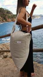 De pie junto al mar, ella apoya el Cojín Ceniza sobre el hombro. Tonos gris y blanco que evocan calma y sencillez, reflejando la elegancia relajada del Mediterráneo.