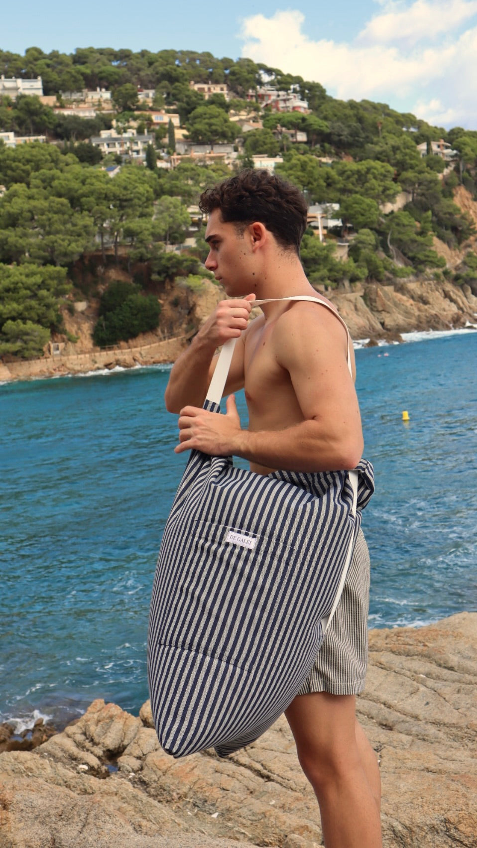 Sobre las rocas junto al mar, él lleva el Cojín Costa al hombro. Azul marino y blanco que reflejan la esencia del Mediterráneo: serenidad, estilo y aventura bajo el sol.