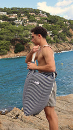 Sobre las rocas junto al mar, él lleva el Cojín Costa al hombro. Azul marino y blanco que reflejan la esencia del Mediterráneo: serenidad, estilo y aventura bajo el sol.