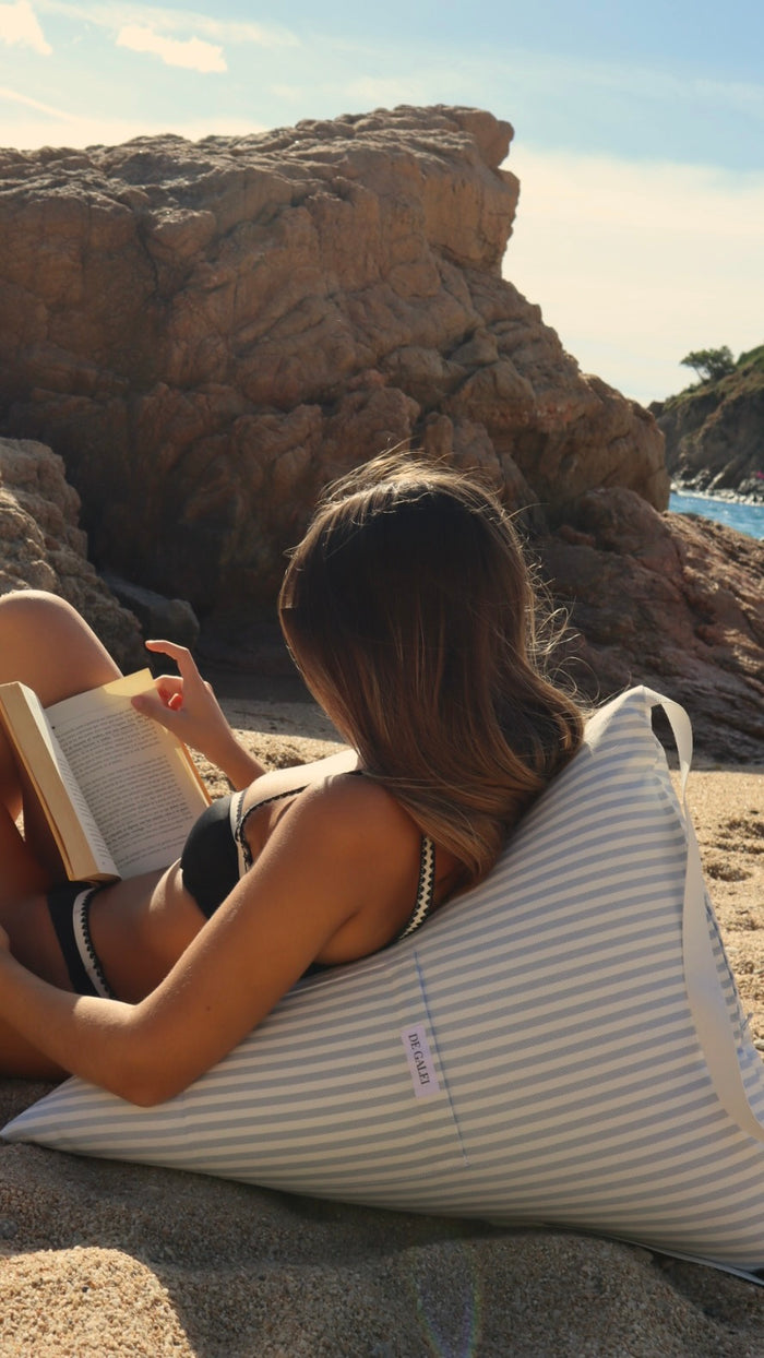 Ella lee bajo el sol sobre el Cojín Creta de De Galei. Azul y blanco que reflejan la calma del mar, creando el escenario perfecto para un momento de descanso y serenidad mediterránea.