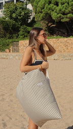 Ella camina sobre la arena con el Cojín Duna al hombro. Un gesto sencillo que resume el verano: comodidad, luz y el inconfundible espíritu mediterráneo de De Galei.