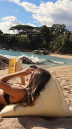Tumbada frente al mar, ella disfruta la calma del verano sobre el Cojín Limón de De Galei. Amarillo y blanco que iluminan la arena, pensados para leer, descansar y sentir el Mediterráneo.
