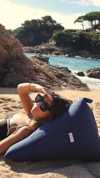 Tumbada sobre la arena, ella descansa sobre el Cojín Marino de De Galei. Azul profundo que evoca el mar, pensado para disfrutar el sol con elegancia y comodidad natural.