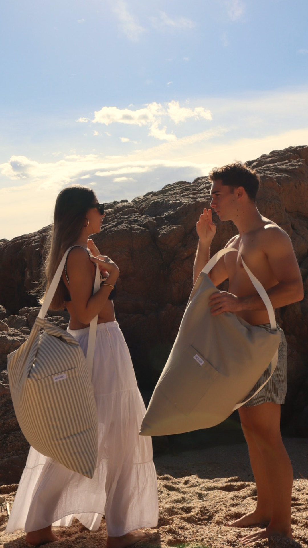 De pie frente al mar, ella lleva el Cojín Duna y él el Cojín Madeira. Tonos beige y blanco que se funden con la arena, reflejando la serenidad y el estilo atemporal de De Galei.